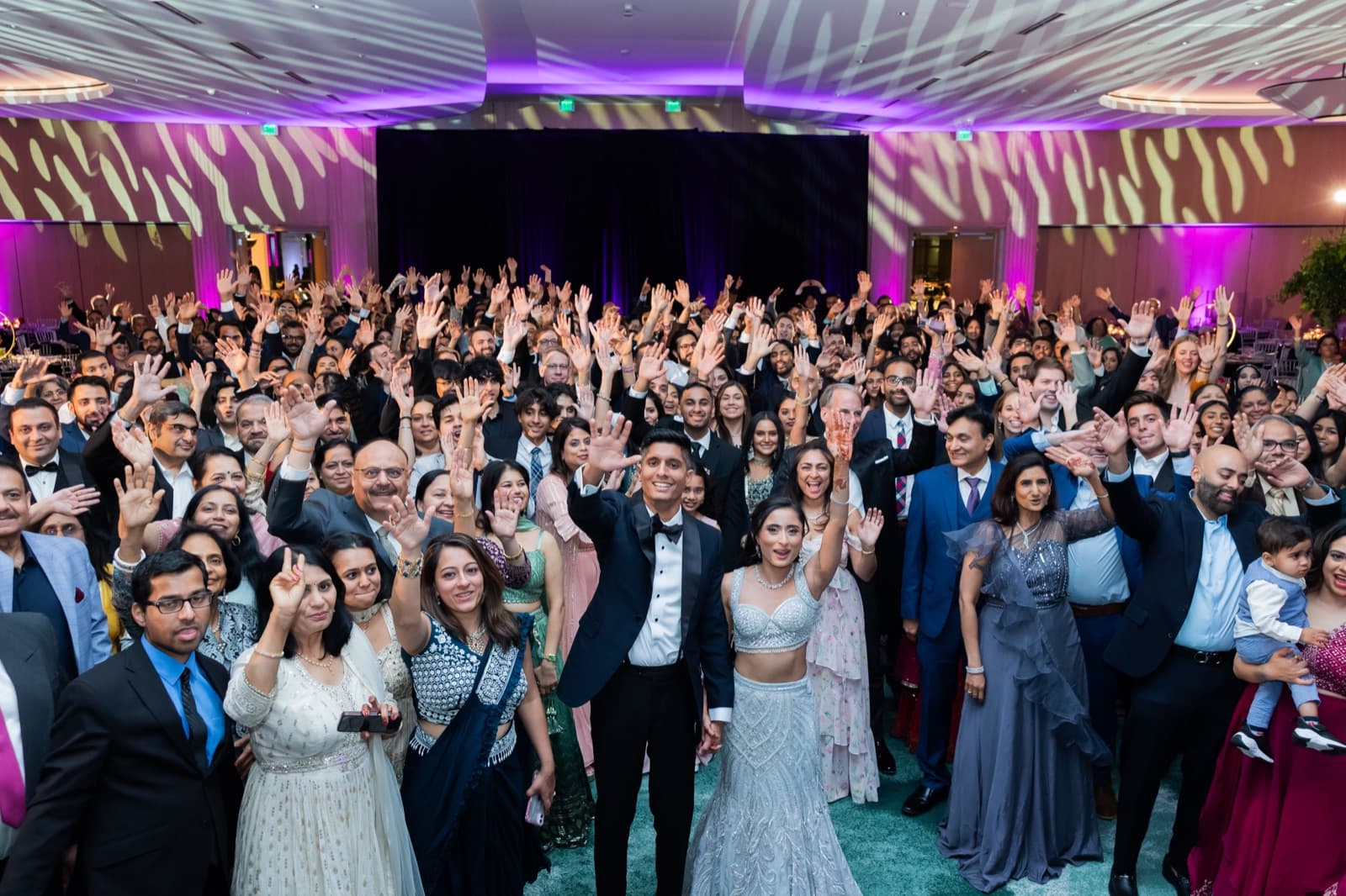 Full wedding party and guests waving for a group photograph under purple and gold patterned lighting