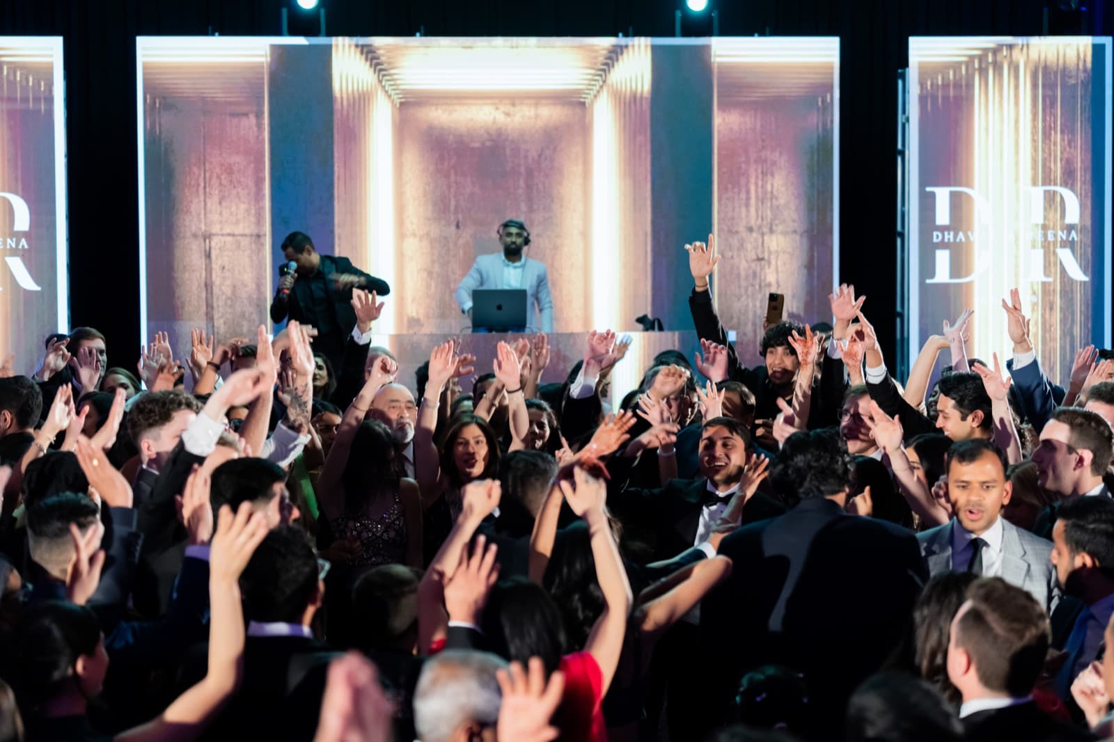 Packed dance floor with hands in the air as the DJ works an elevated booth framed by warm gold LED architecture