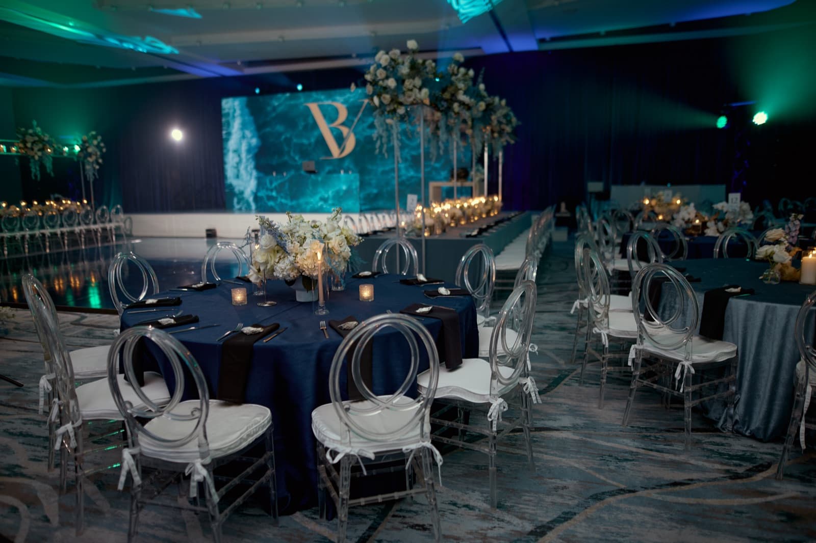 Styled reception room before guests arrive, with navy linens, clear ghost chairs and a teal water-motion LED backdrop